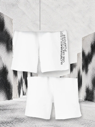 The white Concept bermuda shorts seen from the back, with the lettering on the side and hanging on a thread, and from the front, between white paper walls stained with black.