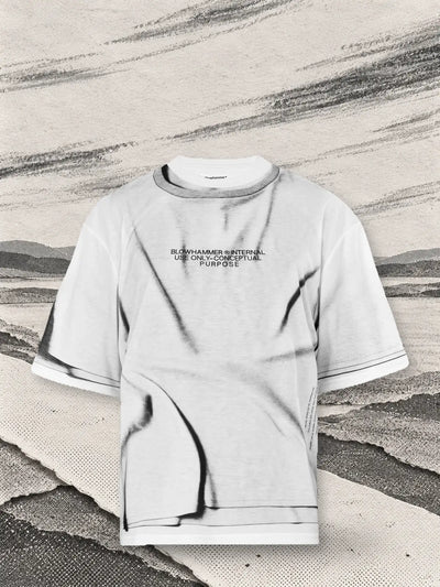 La t-shirt bianca Crux sopra un incrocio di sentieri di carta bianca, con lunghe strisce di carboncino grigio portate dal vento.