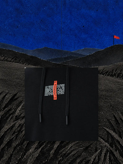 Un pezzo del petto della felpa con cappuccio nera Flag, con l'etichetta rossa, appeso a un filo su una collina di carta nera sotto un cielo blu.