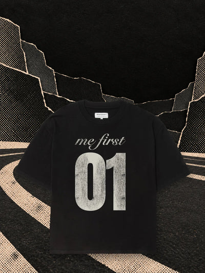 La t-shirt boxy nera MeFirst01 su una pista di carta nera che curva nel buio, accanto a un podio di carta chiara.
