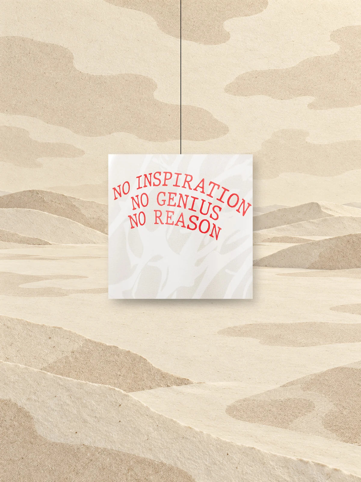 A square cut from the print of the No Genius hoodie, with the red words NO INSPIRATION NO GENIUS NO REASON, hung from a thread above a field of ivory paper.