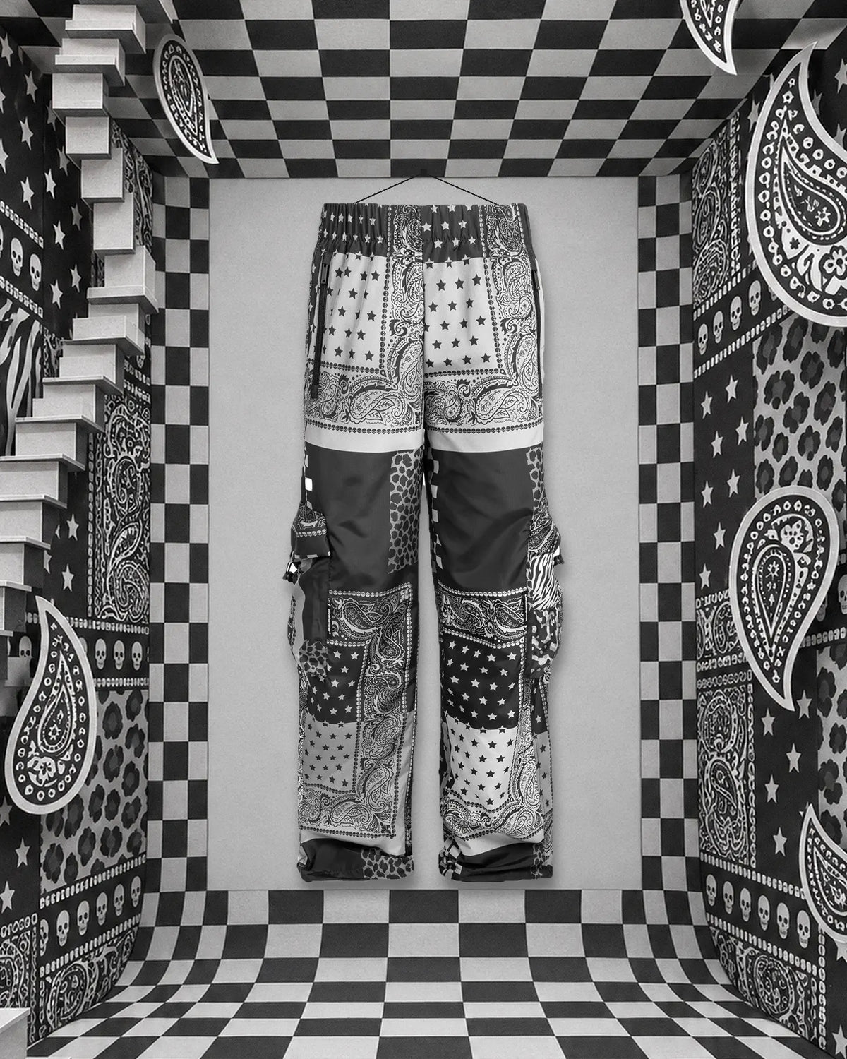 Spodnie cargo Romanticbandana, patchwork czarnych, białych i szarych bandan, zawieszone na nitce na gwoździu wbitym w tylną ścianę papierowego pokoju w białą i czarną szachownicę. Po lewej białe schody, na ścianach paisley, gwiazdy, wzór zebry, cętki i małe czaszki.