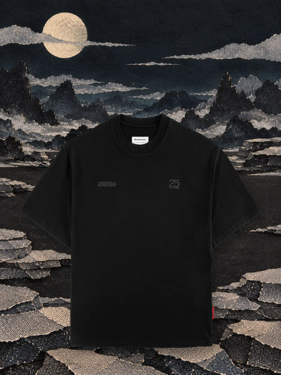 La t-shirt nera Vertebrae su una piana di carta nera di notte, fra file di sassi di carta grigia impilati e la luna.